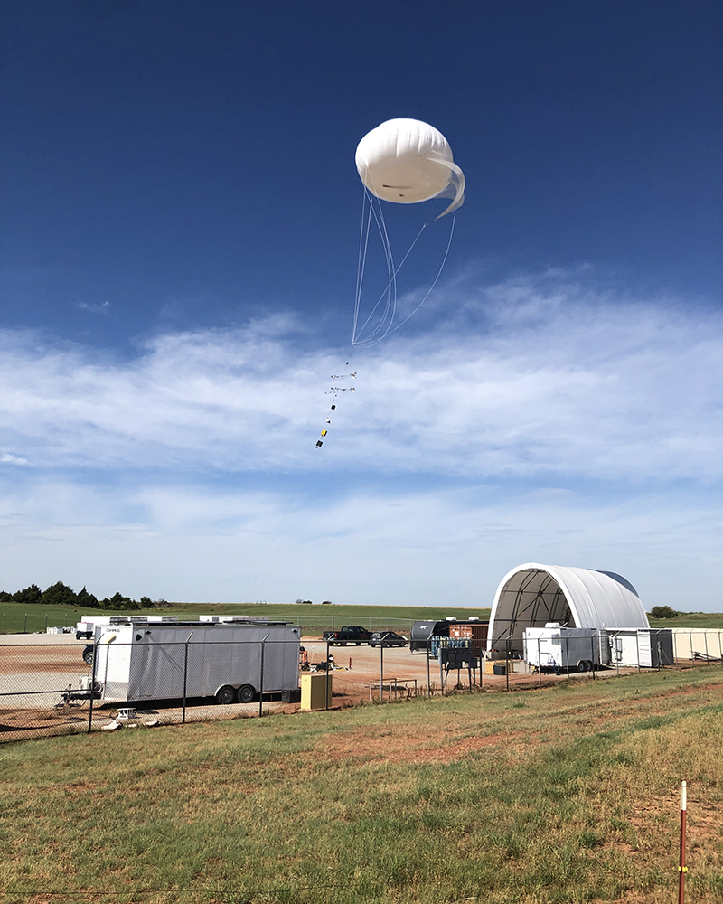 A balloon with instruments on the tether hovers above ARM's Southern Great Plains atmospheric observatory. A trailer and arch-shaped tethered balloon shelter surround the balloon launch area.