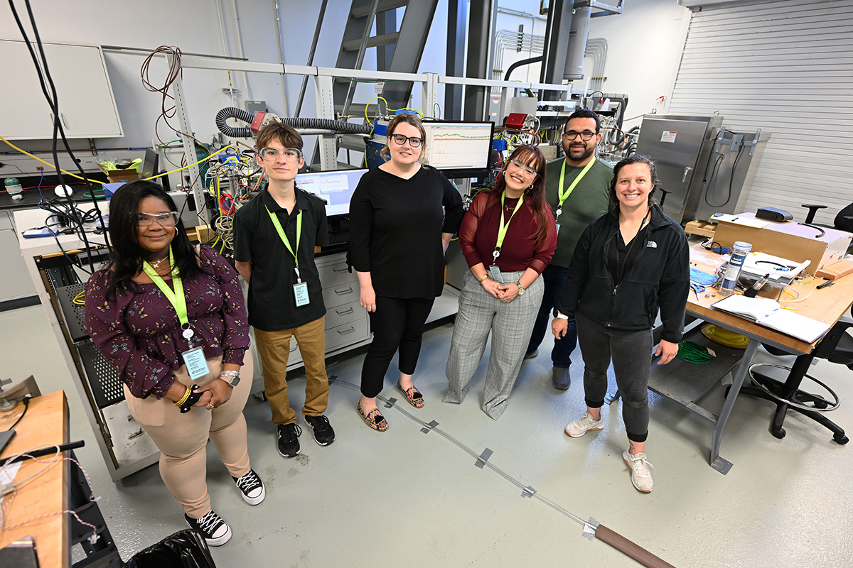 Six people stand in a semicircle. The photo angle is taken from slightly above. The four interns wear eye protection.