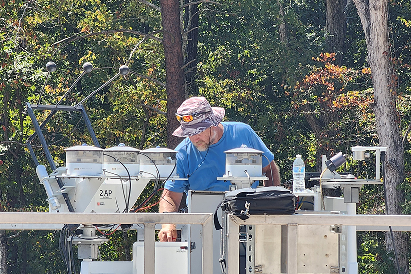 In a forest clearing, a man works with instruments on the roof of an ARM Mobile Facility container.