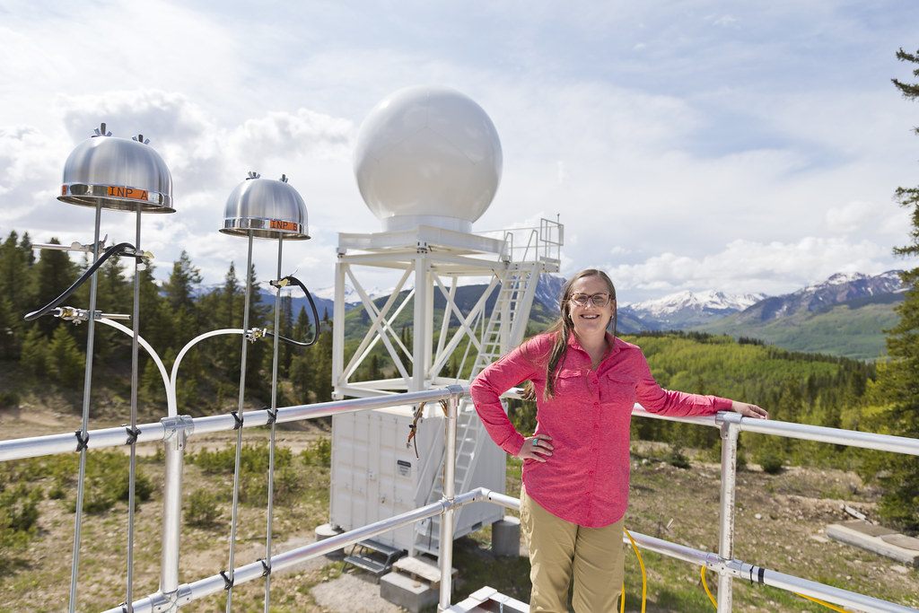 Allison Aiken stands on an instrument platform and rests her left arm on its railing. Two ice-nucleating particle samplers are installed on the railing next to Aiken. Metal salad bowls are flipped upside down to serve as precipitation shields and protect the particle filters underneath. An X-band radar is behind Aiken.