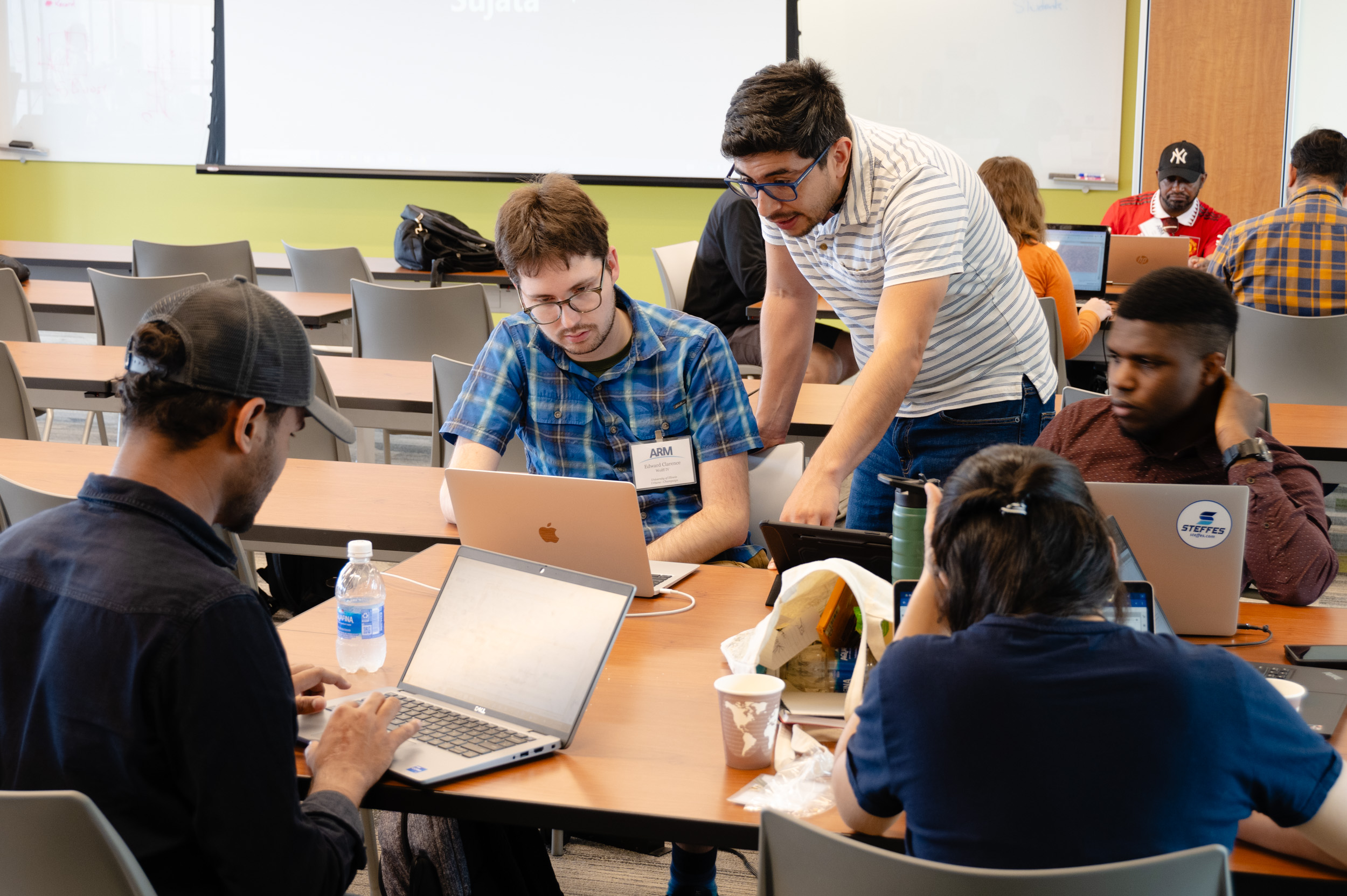 A person standing up leans over to help a group of four people working at a table with laptops open in front of them.