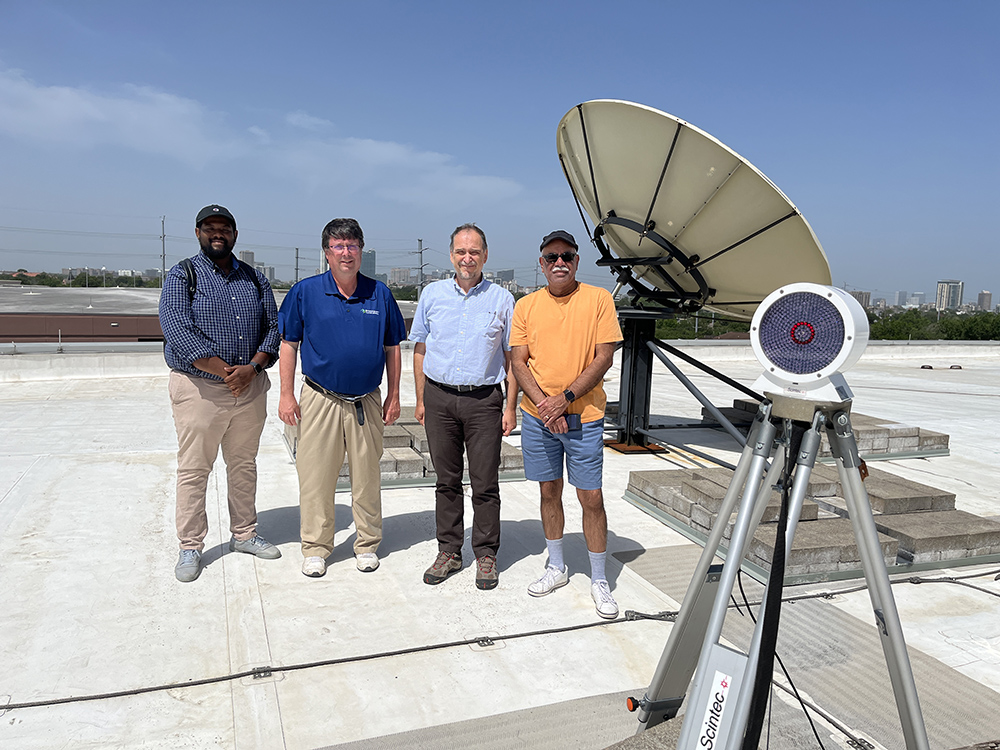 A group of four men stand next to a giant satellite dish on the roof of a building with a downtown skyline in the distance.