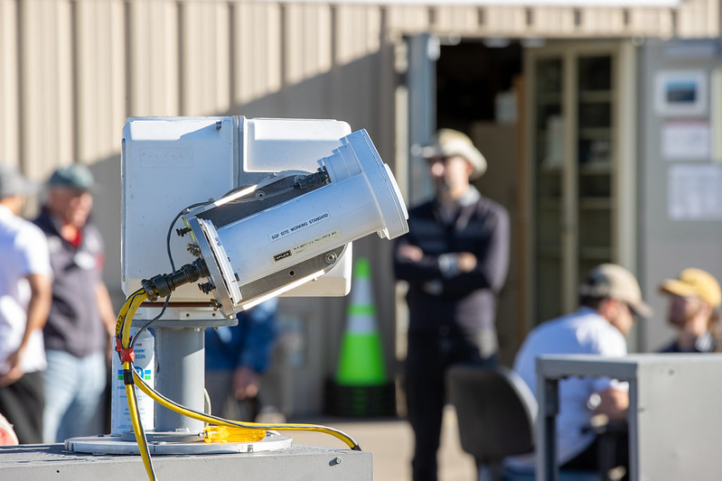 This photo shows the side of a solar tracker and cavity pyrheliometers. A label on the pyrheliometer says "SGP Site Working Standard." People are standing and sitting in the background.