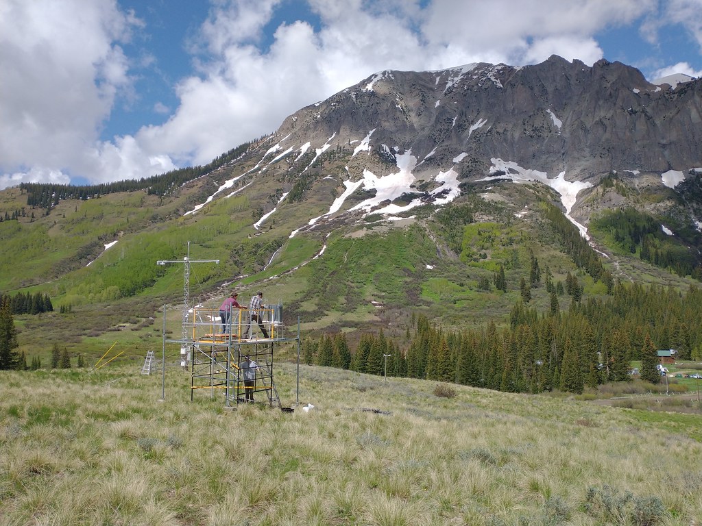 Three people work to break down an instrument platform on a hill overlooking a mountain with some snow lingering in its crevices. A band of trees separates the hill and the mountain. Broken clouds appear behind the mountain.