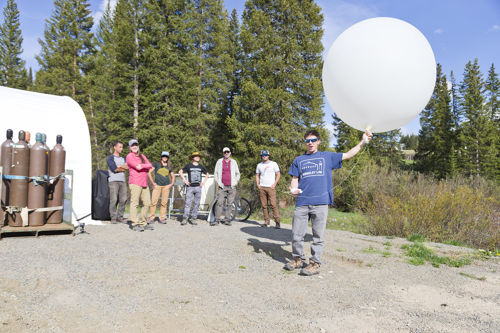 On a patch of gravel in front of tall trees, Daniel Feldman holds the bottom of an inflated balloon in one hand and a radiosonde in the other. Standing behind Feldman are six people awaiting the balloon launch.