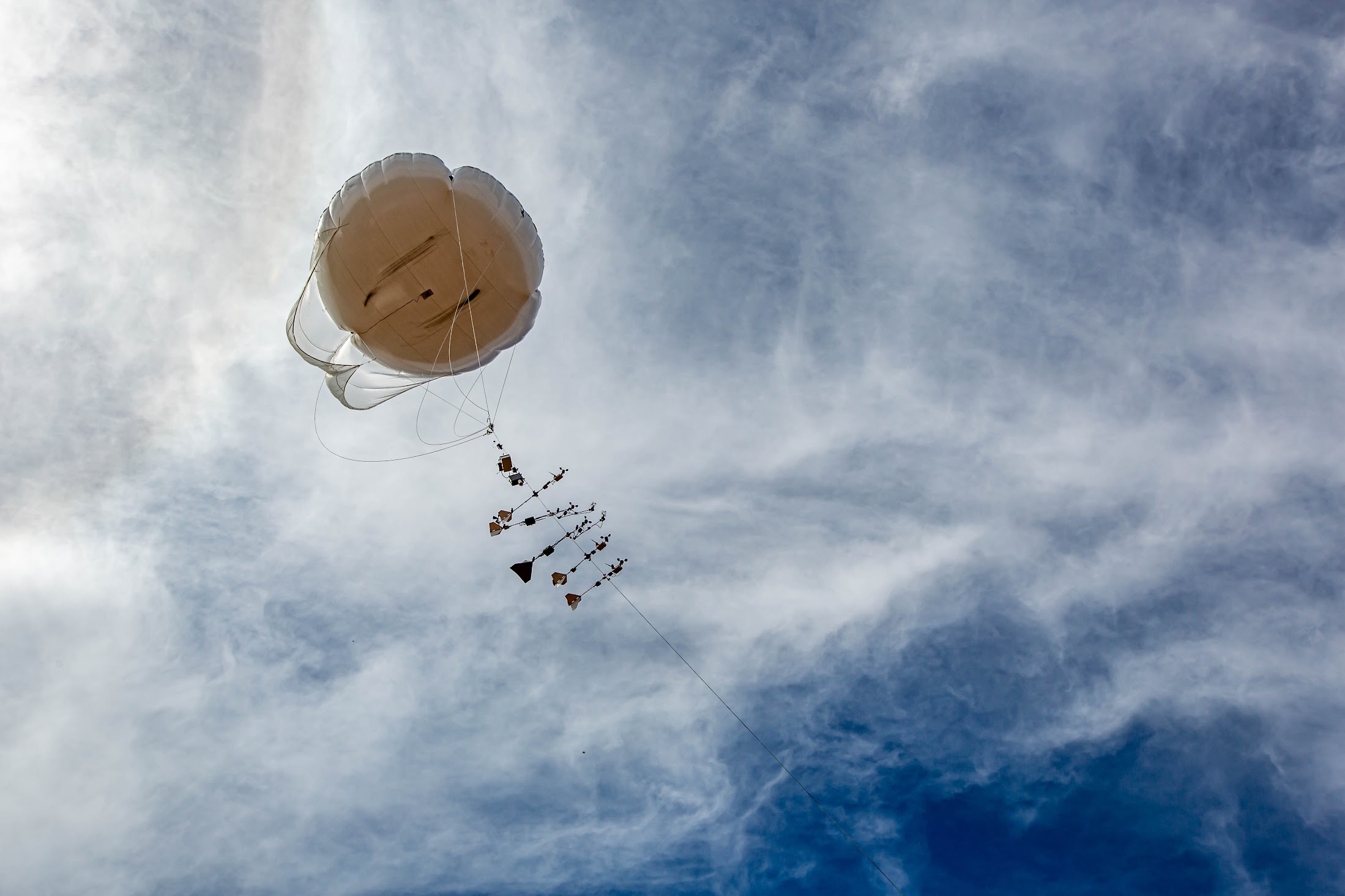 Looking up at swirling clouds, the camera captures a tethered balloon with instruments strung along the tether.