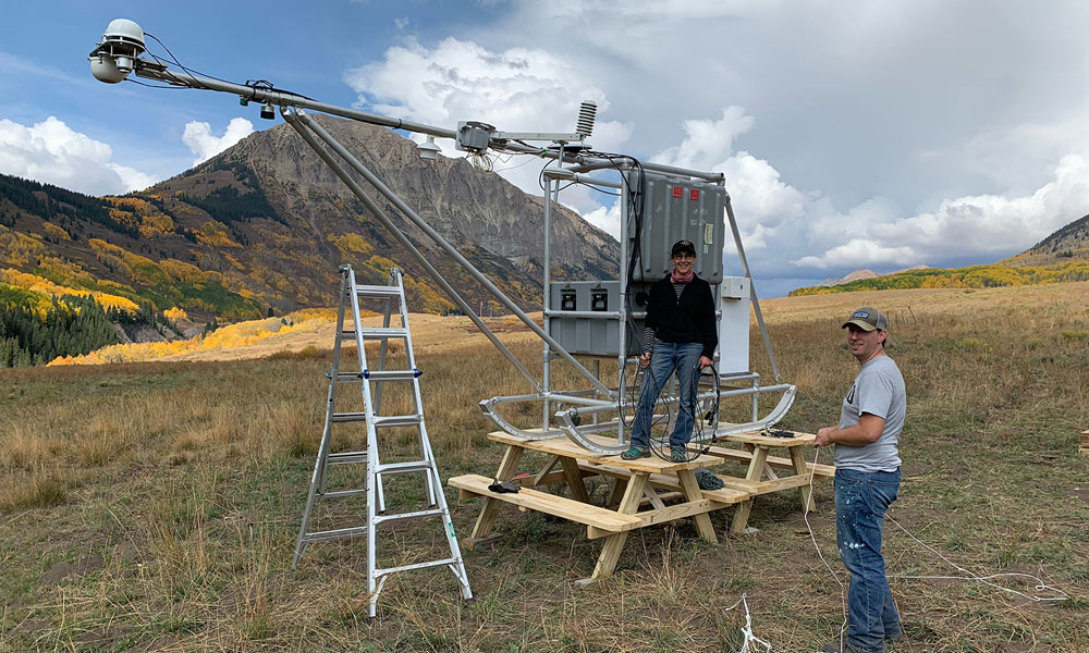 Janet Intrieri stands on a wooden workbench and Christopher Cox on flat ground while posing for a photo before returning to work on their instrument.