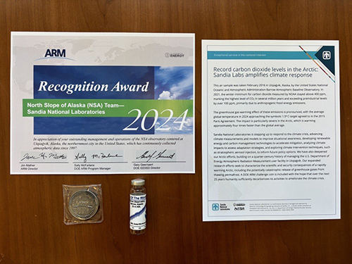 An ARM award certificate, ARM challenge coin, and NOAA air sample are displayed alongside an article detailing what the items represent to the history of Sandia National Laboratories.