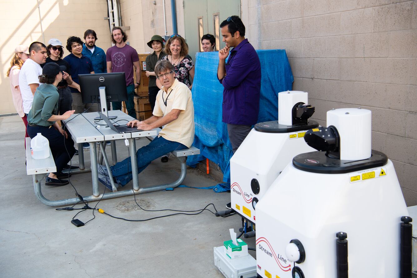 Rob Newsom sits at a laptop while looking at lidars to his left. Raghavendra Krishnamurthy looks down at Newsom and smiles while holding his chin. Lexie Goldberger, sitting across from Newsom, talks to a group of students and their professor looking on. 