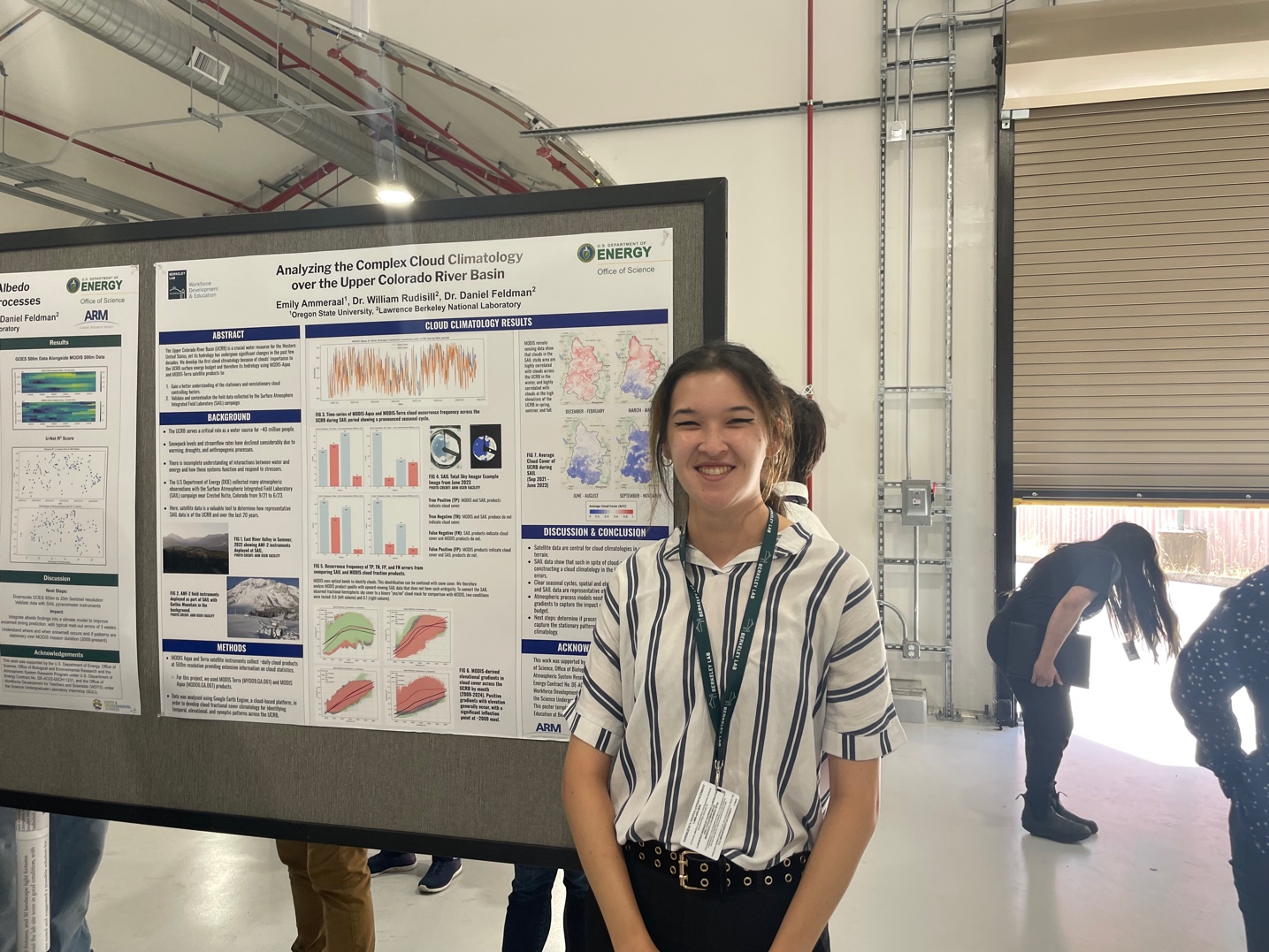 A woman stands next to a scientific poster pinned onto a board. The poster includes, text, data plots, and pictures. The poster says, "Analyzing the Complex Cloud Climatology over the Upper Colorado River Basin."