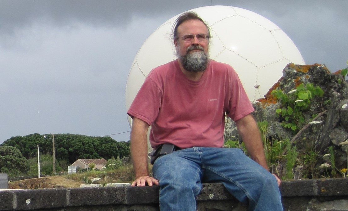 Edward Luke sits on a brick wall with dark clouds behind him.